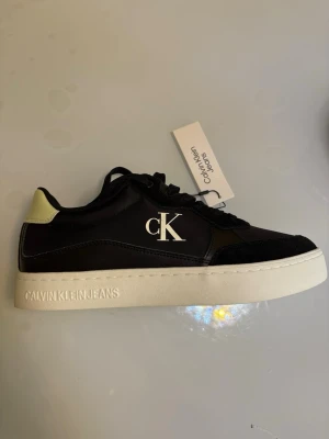 Svarta Calvin Klein Jeans sneakers - Stilrena svarta sneakers från Calvin Klein Jeans med vit sula och kontrasterande ljus detalj vid hälen. Skorna har tydlig CK-logga på sidan och är tillverkade i en mix av läder och textil. Perfekta för dig som gillar klassisk streetstyle.