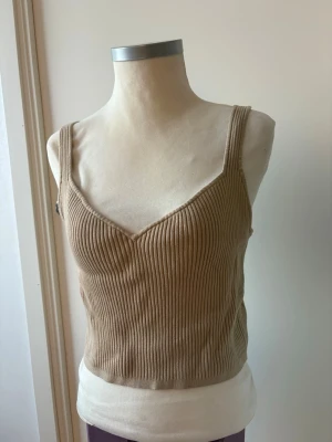 Beige ribbad linnetopp - Snygg beige ribbad linnetopp med smala axelband och v-ringad hals. Toppen är croppad och har en figurnära passform som passar perfekt till både jeans och kjol. Tillverkad i mjukt stickat material för en bekväm känsla.