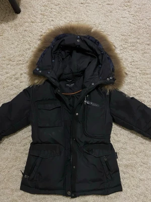 Svart jacka med äkta päls från Hollies - Snygg svart parkasjacka från Hollies med avtagbar huva och äkta päls. Jackan har flera praktiska fickor med dragkedja och tryckknappar, samt en patch med metallogo på ärmen. Perfekt för kalla dagar och riktigt varm.