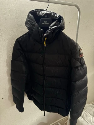 Svart dunjacka från Parajumpers - Snygg svart dunjacka från Parajumpers, modell Sheen. Jackan har huva, dragkedja framtill och den klassiska Parajumpers-loggan på ärmen. Puffig design med breda kanaler och detaljer i gult och orange. Perfekt för kalla vinterdagar.