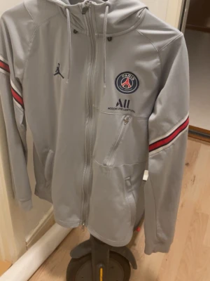 Grå PSG x Jordan zip-hoodie - Snygg grå zip-hoodie från Jordan i samarbete med Paris Saint-Germain. Tröjan har PSG-logga, Jordan-logga och Accor Live Limitless-tryck på bröstet samt röda och svarta ränder på ärmarna. Utrustad med huva och dragkedja framtill.