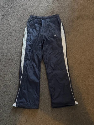 Marinblå byxor från Nike - Säljer ett par marinblå träningsbyxor från Nike med ljusblå sidor längs benen och en liten vit logga på framsidan. Byxorna har elastisk midja, perfekta för träning eller sportiga outfits.