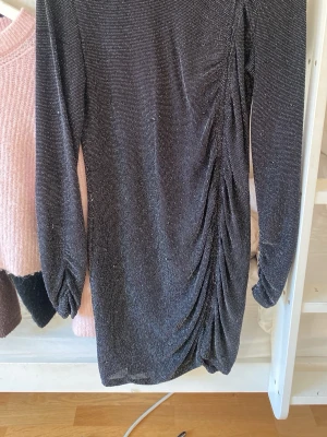 Glittrig svart kortklänning - Snygg svart kortklänning med glittrigt tyg och rynkade detaljer längs ena sidan. Klänningen har långa ärmar och en figurnära passform som ger en trendig look. Perfekt för dig som vill sticka ut med lite extra glitter.