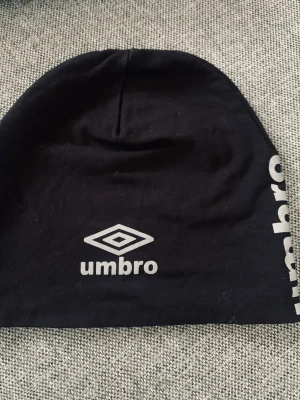Svart mössa från Umbro - Snygg svart mössa från Umbro med vit logga framtill och text längs sidan. Tillverkad i mjukt och stretchigt material som sitter skönt på huvudet. Perfekt för dig som gillar sportig stil och vill ha en enkel men stilren accessoar.