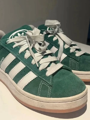 Gröna Adidas Campus sneakers - Gröna Adidas Campus sneakers i mocka med klassiska vita ränder på sidorna och vit sula. De är använda och är lite smutsiga på den vita kanten, men det borde gå att tvätta. 