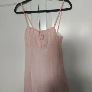Rutig rosa kortklänning från Divided - Supersöt rosa kortklänning från Divided med tunna axelband, justerbar passform och liten rosett framtill. Klänningen har ett rutigt mönster, dragkedja i sidan och är perfekt för varma dagar. Lätt och luftig känsla.