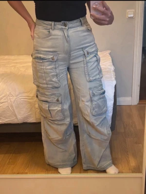 Ljusgrå cargobyxor med vida ben - Säljer ett par ljusgrå cargojeans med flera stora fickor och vida ben. Byxorna har hög midja, knapp- och dragkedjestängning samt detaljerade sömmar. Perfekta för en avslappnad streetwear-look. Kolla min profil för fler plagg! 🥰