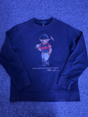 Mörkblå Polo Bear sweatshirt Ralph Lauren - Snygg mörkblå sweatshirt från Ralph Lauren med ikoniska Polo Bear-trycket på bröstet. Tröjan har rund hals, långa ärmar och ribbade muddar. Perfekt för dig som gillar klassisk och sportig stil.