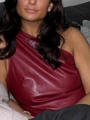 Vinröd one shoulder skinnimitation topp - Snygg vinröd topp i skinnimitation med one shoulder-design. Toppen är ärmlös och har en modern, asymmetrisk skärning som ger en edgy look. Perfekt för dig som vill sticka ut med en trendig och stilren topp.