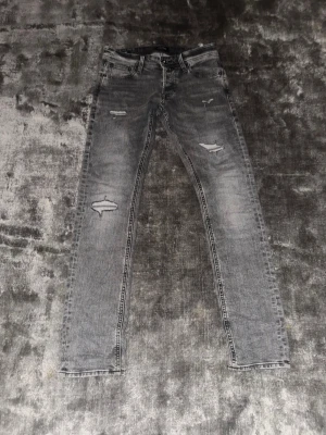Jack & Jones Jeans - Säljer ett par grå jeans från Jack & Jones med slitningar och distressed detaljer på benen. Jeansen är ”slim glenn” och har en klassisk femficksdesign. Perfekta för en avslappnad och trendig look.