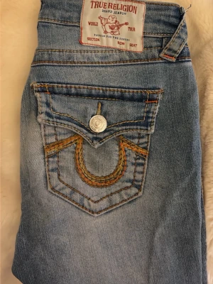 True Religion Joey Low Rise Flare Jeans - Säljer ett par True Religion Joey jeans i ljusblå tvätt med orange kontrastsömmar och snygga broderade bakfickor. Modellen är low rise med flare-ben och  silverknappar. ordinarie pris 1600