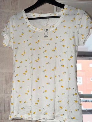 Pyjamas med citronmönster - Vit pyjamas topp med citronmönster. Provad. Toppen har rund halsringning och korta ärmar. Storlek S men passar bäst på XS för avslappnad passform. Tillverkad i 95% viskos 5% elastan. Originalpris: 100kr Nypris: 40kr