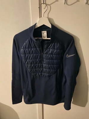Mörkblå Nike tröja med quiltad front - Snygg mörkblå tröja från Nike med quiltad panel framtill och halv dragkedja. Tröjan har lång ärm, hög krage och en diskret Nike-logga på ärmen. Perfekt för lager-på-lager och sportig stil.