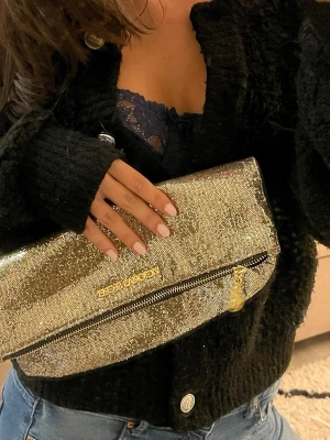 Guldig kuvertväska från Victorias secret - Säljer en glittrig guldig kuvertväska. Väskan har en vikbar design, dragkedja framtill och är täckt av små glittrande paljetter. Perfekt för att lyfta vilken outfit som helst med extra bling. Märkeslogga i guld på framsidan.