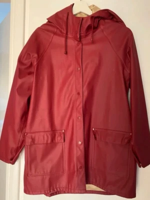 Vinröd regnjacka - Snygg vinröd regnjacka med huva och dragsko. Jackan har tryckknappar framtill och två stora fickor med lock. Tillverkad i vattenavvisande material, perfekt för regniga dagar. Enkel och stilren design som passar till det mesta.