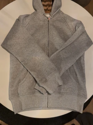 Grå zip hoodie från Burberry - Säljer en grå hoodie från Burberry med dragkedja och klassisk huva. Insidan av huvan har det ikoniska Burberry-mönstret och bröstet pryds av diskret broderad logga. Tillverkad i mjuk bomullsblandning och har ribbade muddar samt fickor framtill.