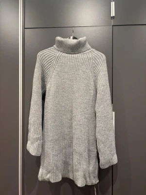 Grå polotröja i ribbstickad modell - Mysig grå stickad polotröja i längre modell med ribbstickat mönster och långa ärmar. Perfekt för lager på lager och har en hög krage som håller dig varm. Enkel och stilren design som passar till det mesta.