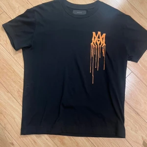 Svart t-shirt från Amiri med orange tryck - Cool svart t-shirt från Amiri med ett orange, droppande logotryck på bröstet. Klassisk rund halsringning och korta ärmar. Perfekt för dig som gillar streetwear och vill sticka ut med en unik detalj.