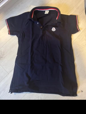 Mörkblå Moncler pikétröja - Snygg mörkblå pikétröja från Moncler med klassisk krage och knappar framtill. Tricolore-detaljer i rött, vitt och blått på krage och ärmslut samt Moncler-logga på bröstet. Tillverkad i 100% bomull för en skön känsla.