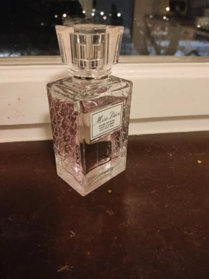 Miss Dior Silky Body Mist parfym - Miss Dior Silky Body Mist från Dior i en elegant, fyrkantig glasflaska med vacker struktur på sidorna. Parfymen har en ljusrosa ton och flaskan har en genomskinlig, stilren kork. Perfekt för dig som gillar lyxiga och feminina dofter.