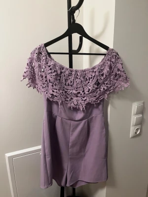 Lila off shoulder jumpsuit AMISU - Supersnygg lila jumpsuit från AMISU i storlek 38. Off shoulder-modell med bred virkad spets upptill som ger en romantisk vibe. Kort byxdel och mjukt tyg som känns skönt mot huden. Perfekt för dig som vill sticka ut med en unik och trendig look.