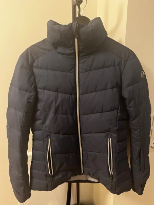 Mörkblå skid-/vinterjacka med fuskpäls - Snygg mörkblå skid-/vinterjacka med hög krage och avtagbar huva med fuskpäls. Jackan har vita dragkedjor framtill och på fickorna samt ett diskret märke på ärmen. Perfekt för kalla vinterdagar och riktigt skön att bära.