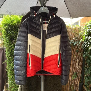 Moncler dunjacka med färgblock - Snygg dunjacka från Moncler med coolt färgblock i mörkblått, vitt och rött. Jackan har huva med snörning, två dragkedjefickor och quiltad design. Perfekt för dig som vill sticka ut med en sportig och modern look. Passar dig som gillar streetstyle.