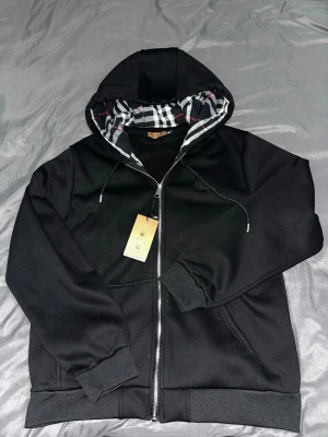 Svart hoodie från Burberry med dragkedja - Svart hoodie från Burberry i fint skick. Rutmönster på insidan av huvan och liten logga på bröstet. Storlek S men passar även M. Skön och stilren
