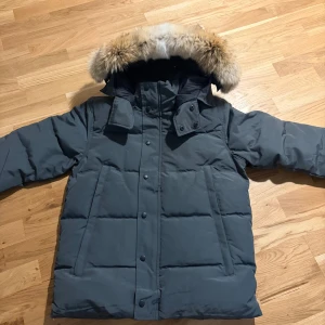 Grå dunjacka från Canada Goose - Snygg grå dunjacka från Canada Goose med avtagbar huva och äkta pälsdetalj. Jackan har en klassisk puffer-modell, svarta knappar och dragkedja samt logotyp på ärmen. Perfekt för kalla vinterdagar.