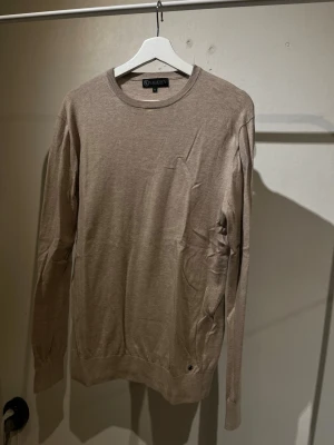 Beige stickad tröja - Snygg beige stickad tröja från Lagency (Therlunds). med rund halsringning och långa ärmar. Tröjan har en enkel och stilren design med ribbade muddar vid ärmslut och nederkant. Perfekt för dig som gillar en clean och klassisk look.