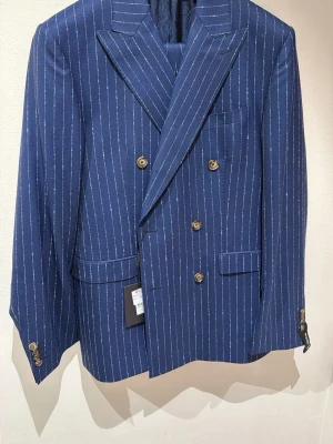Blå randig kostym från Sand - Snygg blå dubbelknäppt kostym från Sand Copenhagen med vita kritstrecksränder och modern fit. Kavajen har breda slag, fyra knappar vid ärmslutet och flera fickor framtill. Perfekt för en stilren och trendig look.