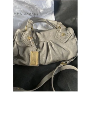 Grå handväska från Marc Jacobs - Snygg grå handväska från Marc Jacobs i mjukt läder med guldfärgade metalldetaljer och dekorativ bricka framtill. Väskan har både handtag och axelrem för flexibel användning. Klassisk design med fina sömmar och detaljer.