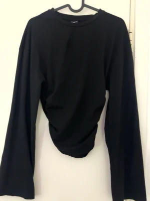 Svart långärmad topp med rynk - Svart långärmad topp med rund hals och snygg rynkdetalj på ena sidan. Toppens material känns mjukt och stretchig vid midjan