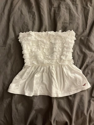 Vit volang bandeau topp Hollister - Supersöt vit bandeau topp från Hollister med volanger upptill och utsvängd nederdel. Toppens material känns mjukt och lätt, perfekt för varma dagar. Liten röd Hollister-logga broderad nertill. Snygg att styla med jeans eller kjol! Är i storlek m men passar på mig som brukar ha xxs/xs
