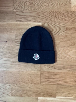 Svart Moncler mössa med logotyp - Snygg svart mössa från Moncler med klassisk uppvikt kant och broderad logotyp framtill. Mössan är ribbstickad och har en enkel, stilren design som passar perfekt till kalla dagar. Materialet känns mjukt och värmande.