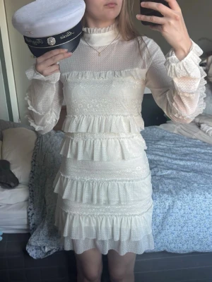 Vit volangklänning - studentklänning  - Supersöt vit kortklänning med volanger och spetsdetaljer. Klänningen har hög hals, långärmade ärmar med volangavslut och genomgående romantiska spetsinslag. 