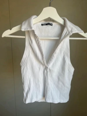 Vit ribbad topp med krage från Zara - Snygg vit ribbad linnetopp från Zara med krage och djup v-ringning. Toppen är ärmlös och croppad, perfekt för en trendig och avslappnad look. Passar till både jeans och kjol för en cool stil. 93% bomull 7% elestan. 