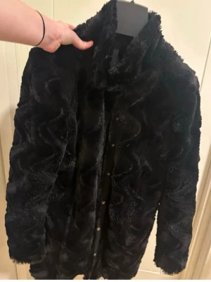 Svart fuskpälsjacka med mönster stl L  - Snygg svart pälsjacka i fuskpäls med vågigt mönster och knappar framtill. Jackan har hög krage och är perfekt för kalla dagar. Den är mjuk och har en trendig look som passar till många outfits. Fint begagnat skick :)