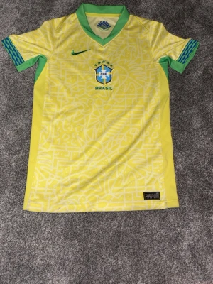 Brasilien matchtröja 24/25 från nike - Säljer en officiell Brasilien matchtröja från Nike år 2024 i gult med gröna detaljer och blått märke. Tröjan har ett grafiskt mönster, korta ärmar och V-ringad krage. Tvättbar liten fläck på backsidan av tröjan och använd 3 gånger. OBS den är i BARN M inte man M
