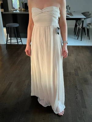 Ljusbeige axelbandslös långklänning - Elegant axelbandslös långklänning i ljusbeige med twistad detalj vid bysten och lätt, flytande tyg. Klänningen har dragkedja i ryggen och en mjukt veckad kjol som ger ett romantiskt intryck. Perfekt för speciella tillfällen.