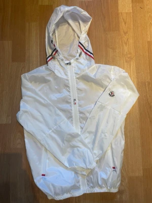 Vit Moncler vindjacka med huva - Snygg vit vindjacka från Moncler med huva och dragkedja. Jackan har Moncler-logga på ärmen och detaljer i rött och blått vid huva och fickor. Lätt och stilren design i glansigt material, perfekt för blåsiga dagar.