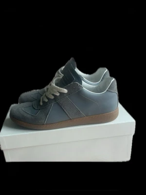 Grå Maison Margiela sneakers - Snygga grå sneakers från Maison Margiela med ovandel i mocka och läder, vita snören och klassisk gummisula. Ikonisk design med diskreta detaljer och stilren look som passar perfekt till streetwear och casual outfits.