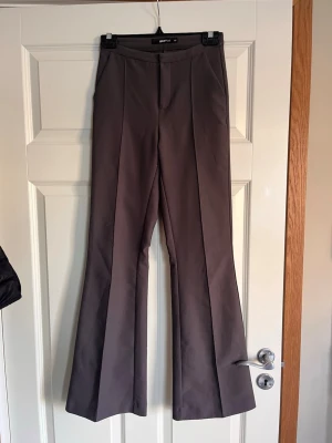 Bruna utsvängda kostymbyxor Gina Tricot - Snygga bruna kostymbyxor från Gina Tricot med hög midja och utsvängda ben. Byxorna har pressveck framtill och en stilren look utan synliga fickor fram. Perfekta för dig som gillar trendiga och klassiska plagg.