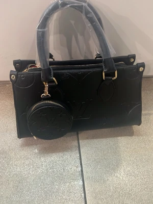 Svart axelremsväska med gulddetaljer - Stilren svart handväska i slätt konstläder med guldfärgade metalldetaljer och avtagbar axelrem. Väskan har en enkel, rektangulär form och stängs med dragkedja. Perfekt för dig som gillar minimalistisk och trendig design.