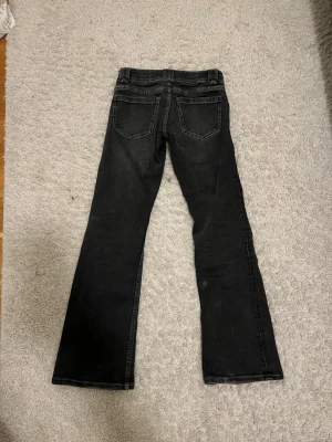 Svarta bootcut jeans med fickdetaljer - Snygga svarta bootcut jeans med dekorativa fickor fram som har lock och knapp. Jeansen har klassisk femficksdesign och subtila slitningar för en cool look. Perfekta för dig som gillar en avslappnad men trendig stil.