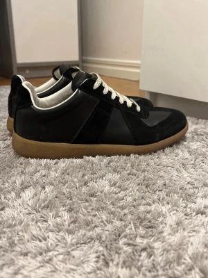 Maison Margiela svarta sneakers - Maison Margiela sneakers i svart läder och mocka med vita snören och klassisk gummisula i beige. Kvitto finns .Låg modell med stilren design och diskreta detaljer.Perfekta för dig som gillar minimalistiskt och trendigt mode. Har även box till skorna.