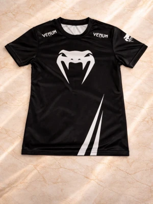 Svart sporttröja från Venum - Snygg svart sporttröja från Venum med vit orm-logga framtill och coola vita detaljer. Stora Venum-tryck på både fram- och baksida. Tillverkad i ett lätt och snabbtorkande material, perfekt för träning eller kampsport.