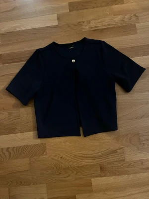 Svart kortärmad kofta med pärlknapp - Stilren svart kofta med korta ärmar och en dekorativ pärlknapp framtill. Modellen är rak och något boxig, vilket ger en modern look. Perfekt att bära över ett linne eller t-shirt. Materialet ser ut att vara mjukt och något tjockare för extra komfort.🥰🥰