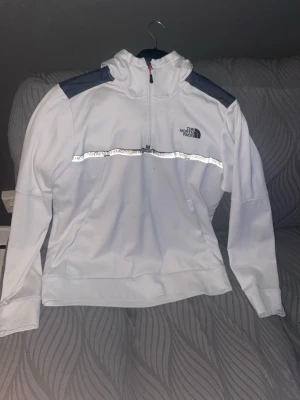 Vit hoodie från The North Face - Snygg vit hoodie från The North Face med mörkgrå axelpartier och reflekterande detalj över bröstet. Halv dragkedja framtill och klassisk huva. Perfekt för dig som gillar streetwear och sportig stil.
