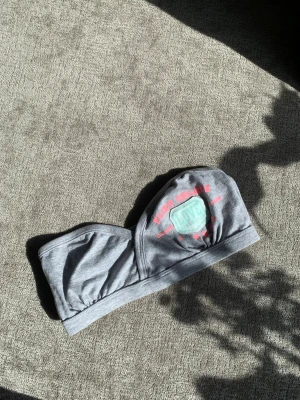 Söt bandeau-bh med tryck 💕 - Säljer en grå bandeau-bh i mjukt bomullsmaterial med tryck i rosa och mintgrönt framtill. Bh:n har en sportig look utan axelband och bred resår nedtill för extra stöd. Perfekt för dig som gillar enkel och bekväm stil med det lilla unika 💕